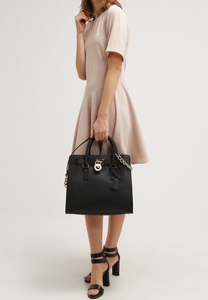 Sac à main structuré noir avec doubles poignées, quincaillerie dorée et un pendentif. Texture lisse et mate ; contraste avec une robe rose clair.