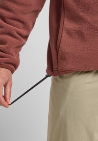 Pull en polaire bordeaux avec des accents côtelés et un cordon de serrage à l'ourlet, associé à un pantalon beige léger. La main tire sur le cordon de serrage.
