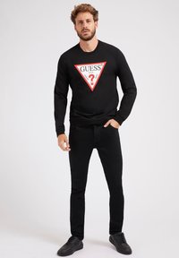 Svart hettegenser med en triangelformet rød og hvit logo som har "GUESS" og et spørsmålstegn i midten, kombinert med svarte bukser og joggesko.