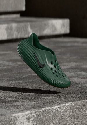 Ciemnozielony but Nike typu slip-on z otworami wentylacyjnymi unoszący się nad betonowymi schodami przy stonowanym oświetleniu.