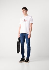 Hvit bomull t-skjorte med "Calvin Klein Jeans" logo, blå denimbukser, svarte sneakers med såle i beige, og en svart skulderveske.