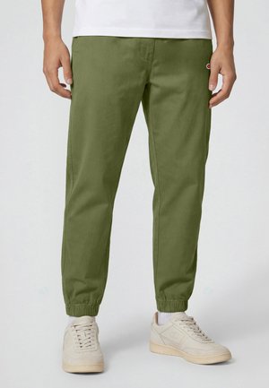 Mężczyzna w oliwkowych spodniach dresowych z elastycznymi mankietami, białych skarpetkach, beżowych sneakersach i białej koszuli, stojący na jasnym tle.