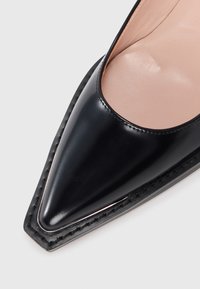 Black leather pointed-toe shoe with a glossy finish, featuring an open side and a smooth inner lining. Stitched detailing along the sole.