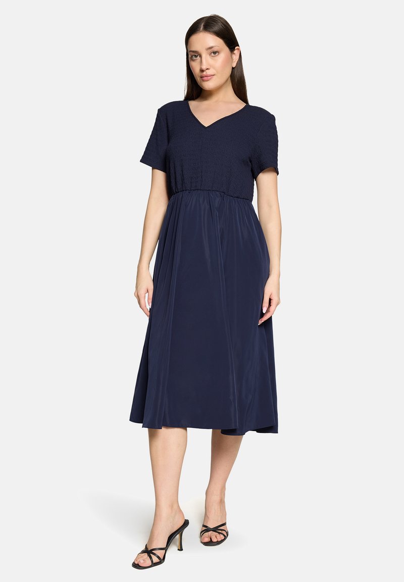 Robe marine avec manches courtes légèrement bouffantes ; corsage froncé menant à une jupe fluide jusqu'aux chevilles ; portée avec des talons noirs à brides.