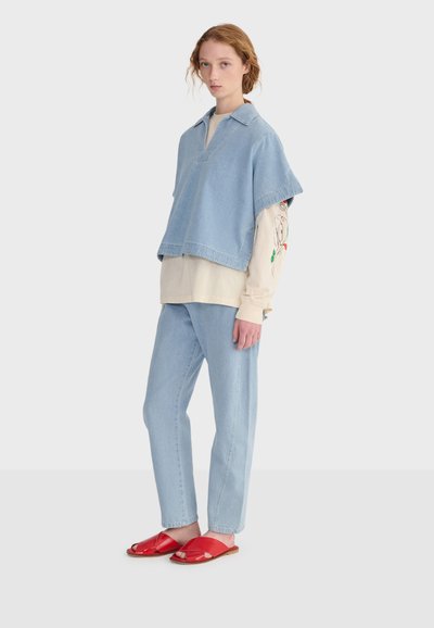 Femme portant un gilet et un pantalon en denim bleu clair, une chemise beige à manches longues avec un motif sur les manches, et des sandales ouvertes rouges, debout devant un fond blanc.
