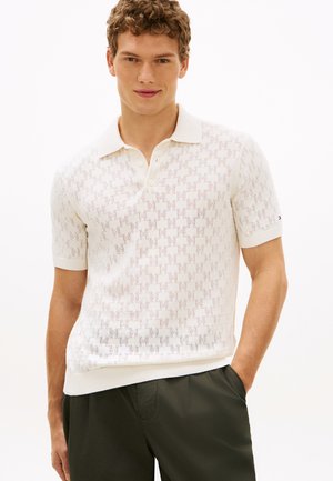 Młody mężczyzna w białej koszulce polo z krótkim rękawem i teksturowanym wzorem w formie krzyża oraz ciemnozielonych spodniach, stoi z jedną ręką w kieszeni.