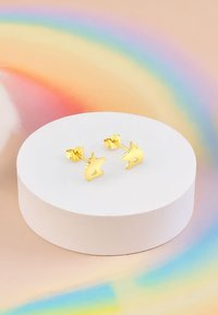 Orecchini a forma di unicorno in oro con dettagli intagliati, esposti su un piedistallo rotondo bianco contro uno sfondo arcobaleno pastello.