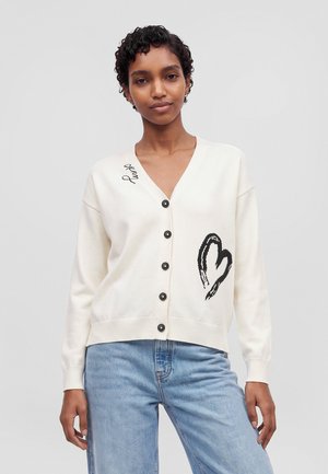 Un modèle porte un cardigan blanc boutonné sur le devant, orné d’un cœur noir, et un jean bleu clair sur un fond uni.