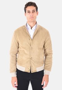 Giovane uomo che indossa una giacca beige in pelle scamosciata con bottoni, polsini e orlo a maglia, camicia bianca e pantaloni neri, in piedi su uno sfondo neutro.