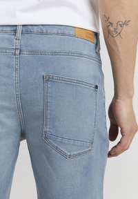 Svijetloplave traperice imaju stražnji džep sa detaljima šivanja. Pojas ima smeđu kožnu oznaku s utisnutim natpisom "DENIM JOURNEY".