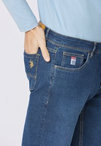 Jeans in denim blu scuro con un logo ricamato in oro sulla tasca posteriore, una finitura testurizzata e dettagli di cucitura visibili.