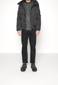 Svart pufferjacka med huva, med en vadderad design och tryckknappar, bärs över en grön mönstrad skjorta och svarta byxor. Svarta sneakers.