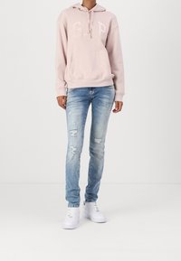 Sudadera con capucha de color rosa claro con el logo de "GAP", combinada con unos vaqueros azul claro desgastados y zapatillas deportivas blancas. Conjunto casual con un ajuste relajado.