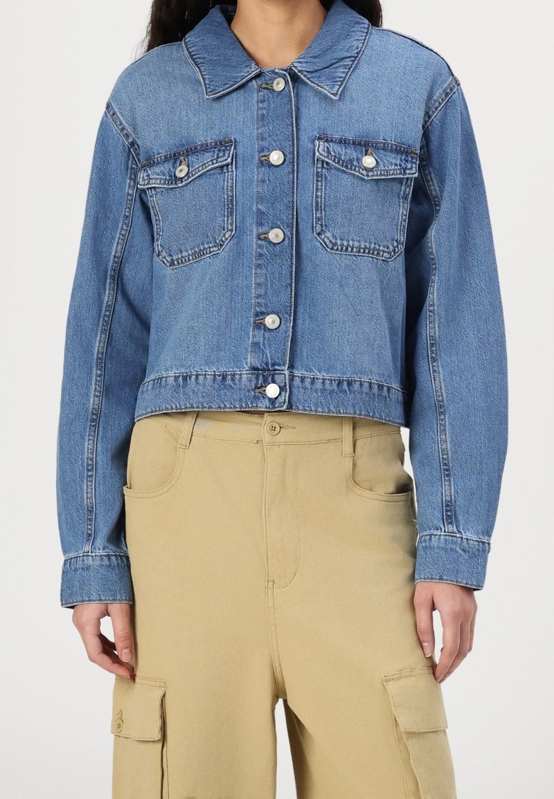 Femme portant une veste courte en jean bleu avec des poches devant et un pantalon cargo beige taille haute avec poches latérales.