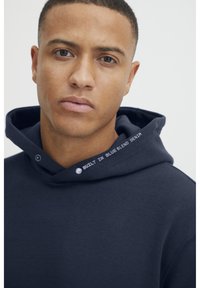 Mornarsko modra kapuca z mehkimi vlakni, z logotipom na ovratniku in nastavljivimi vrvicami na kapuci.