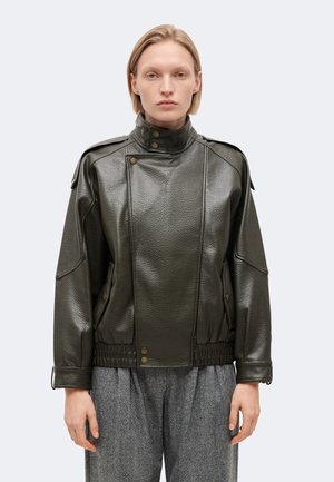 Personne aux cheveux blonds et raides porte une veste en cuir texturée vert foncé avec un col montant et des boutons, associée à un pantalon gris.