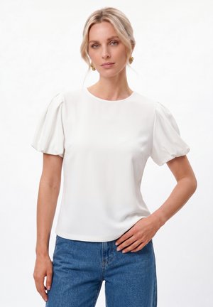 Blonde Frau mit goldenen Ohrringen, einer weißen Bluse mit Puffärmeln und blauen Jeans, steht mit einer Hand in der Hüfte vor weißem Hintergrund.