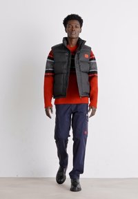 Zwarte puffer vest met hoge kraag gedragen over een rood en zwart gemusterd trui. Donker cargo pants en zwarte schoenen maken de look compleet.