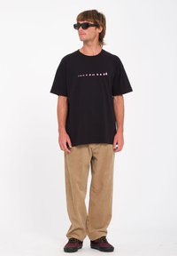Zwarte t-shirt met roze tekst, beige corduroy broek en donkere sneakers. Model draagt zwarte zonnebril en staat tegen een witte achtergrond.
