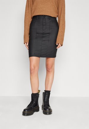 Falda de mezclilla negra con detalles de costura en la parte frontal, combinada con un suéter de punto marrón y botas militares negras con cordones. La falda tiene una silueta ajustada.