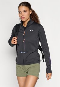 Μαύρο μπουφάν με φερμόρ και ψηλό γιακά, πορτοκαλί φερμουάρ, μωβ και μπλε λεπτομέρειες, φορεμένο πάνω από μαύρη μπλούζα με πράσινο σορτσάκι.