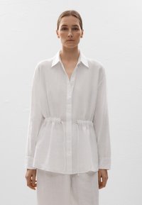 Chemise blanche à manches longues en tissu léger, dotée d'un col de chemise et d'un cordon de serrage réglable à la taille pour un look ajusté.