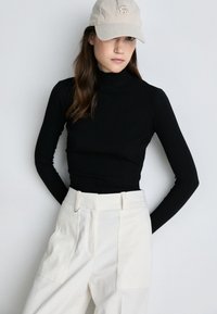 Melna ribota turtlenecka, kas kombinēta ar augsta vidukļa krēmkrāsas biksēm. Modelis valkā gaiši beiju beisbola cepuri ar logotipu.