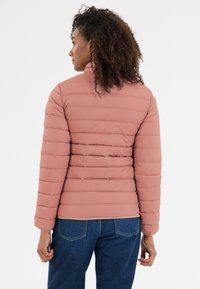 Støvet lyserød pufjakke med lodret quiltet mønster, høj krave og elastiske manchetter. Bæret med blå jeans, der viser en tætsiddende silhuet.