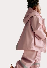 Růžová nepromokavá bunda s kapucí a boční kapsou, doplněná o odpovídající kalhoty. Obsahuje reflexní prvky na rukávech a kalhotách.