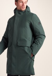 Dark green waterproof parka with a high collar, front zip, and two front pockets. Features a logo on the upper arm. Smooth texture.