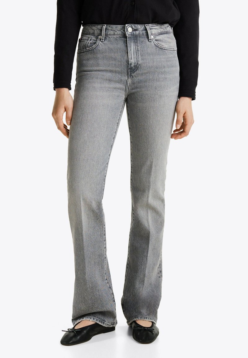 Personne portant un jean gris taille haute droit avec bouton et fermeture éclair, associé à des ballerines noires et un haut noir à manches longues.