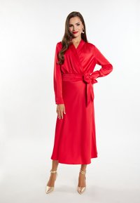 Abito avvolgente in satin rosso con maniche lunghe e vita legata. La gonna è di lunghezza midi ed è abbinata a tacchi con cinturino alla caviglia dorati.