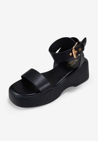 Sandalias de plataforma negras con dos correas de cuero anchas y una correa en el tobillo con una hebilla dorada. Textura suave; diseño ligero.