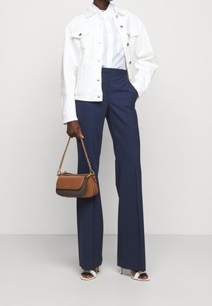 Vrouw draagt een witte jas en blouse, marineblauwe wijde broek met hoge taille, witte sandalen met hak, en houdt een kleine bruine handtas met kettingriem vast.