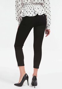 Kvinna som bär svarta tajta byxor, svarta högklackade skor och en vit blus med svarta prickar, sedd bakifrån.