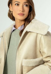 Giacca in shearling beige con colletto crema e tasche frontali, indossata sopra un maglione verde chiaro. Texture liscia con chiusura a zip.