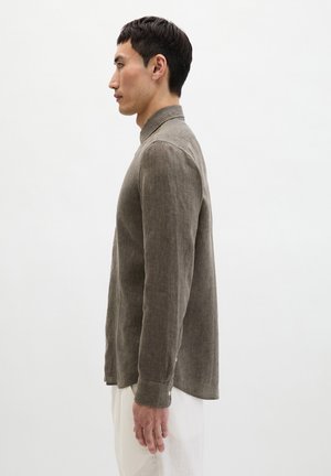 Homme debout de profil portant une chemise en lin marron à manches longues et un pantalon blanc sur un fond blanc uni.