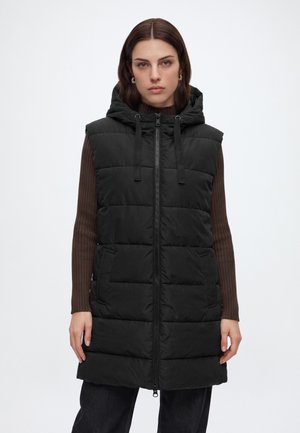 Vrouw draagt een mouwloze zwarte gewatteerde bodywarmer met capuchon en voorrits over een bruine geribbelde trui met lange mouwen.