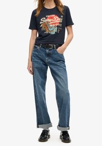 Marinblå t-shirt med vintage-inspirerad grafik och färgglada accenter, tillsammans med ljusblå, vida denimjeans. Utrustad med ett svart bälte och upprullade ärmslut.