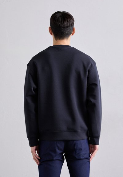 Sweatshirt noir à col rond avec une finition texturée, coupe ample et poignets côtelés, associé à un pantalon bleu marine, vu de dos.