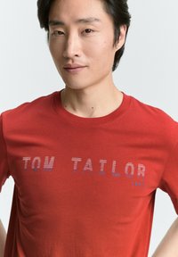 Camiseta de algodón roja con el texto "TOM TAILOR 1962" en rayas azules y blancas. Cuello redondo clásico y mangas cortas. Textura suave.