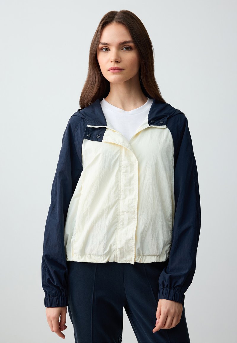 Leichte, zweifarbige Jacke mit navyblauem Oberteil und cremefarbenem Unterteil. Verfügt über eine Kapuze, einen Reißverschluss und elastische Bündchen. Glatte Textur.