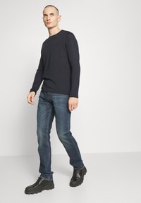 Camicia a maniche lunghe blu navy abbinata a jeans blu e stivali neri robusti. La camicia ha un collo a giro e maniche aderenti, offrendo un look casual.