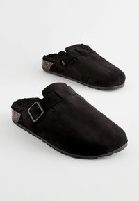 Crni slip-on klompe od teksturiranog antilopa s remenom na kopču. Potplat ima svijetlu mrljavu akcentu za kontrast.