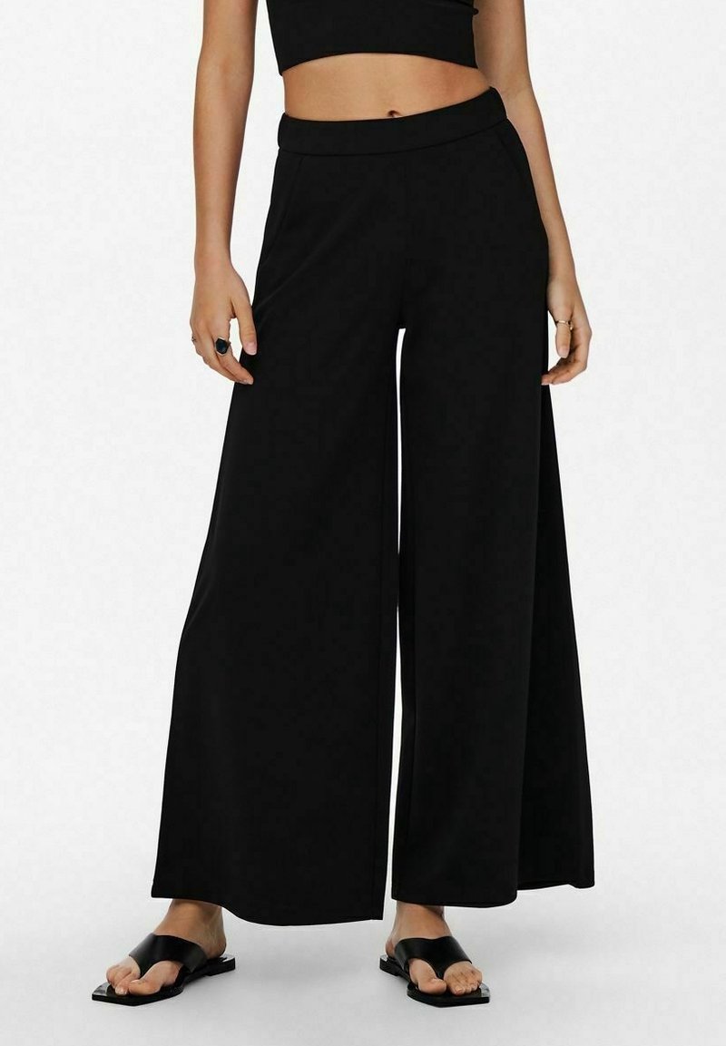 Femme portant un pantalon large noir, un crop top noir, des sandales noires, se tenant debout avec les mains détendues sur le côté sur un fond blanc.