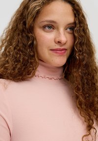 Aansluitende, lichtroze coltrui met ribbeltextuur en een ruchesrand rond de halslijn. Krullend haar gedeeltelijk zichtbaar.