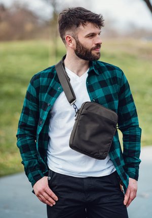 Braune Umhängetasche mit einem schlanken Design, aus weichem Material gefertigt, mit einem Reißverschluss und verstellbarem Tragegurt, getragen über einem karierten Hemd.