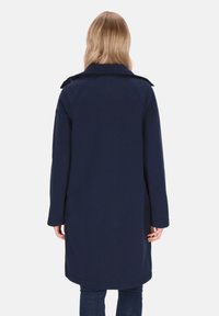 Cappotto lungo blu navy con design dritto, presenta un colletto, maniche lunghe e un tessuto liscio e strutturato. La pulizia è senza decorazioni.