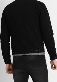 Person med en svart denimjacka och svarta jeans, sedd bakifrån, mot en enkel ljus bakgrund.