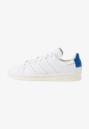 Biały sneaker ze sznurowadłami, perforowane paski po bokach, niebieska naszywka na pięcie i kremowa podeszwa z logo Adidas z boku.